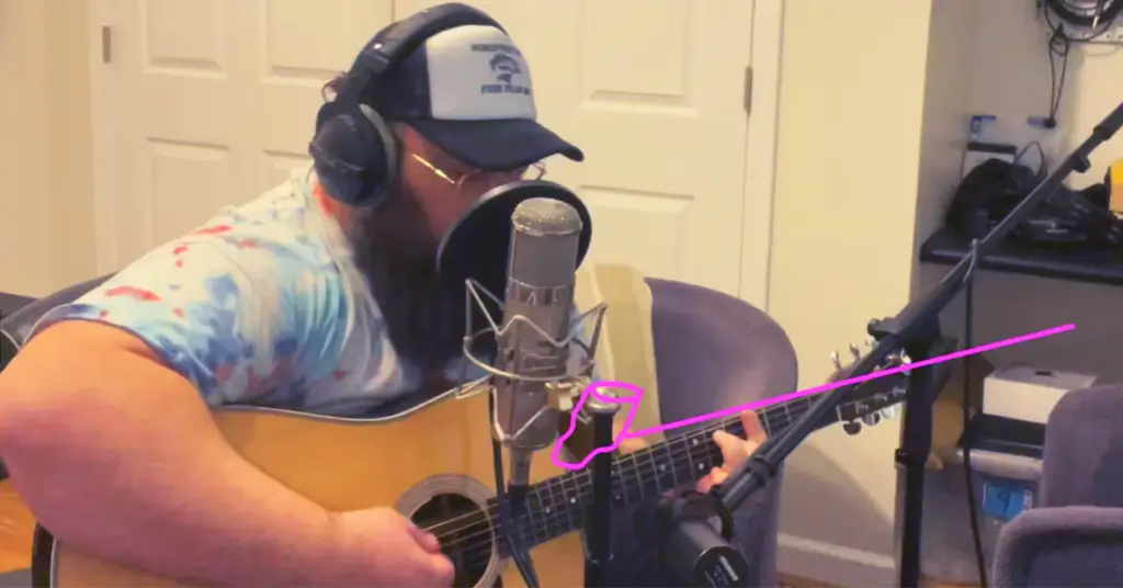 Image and pink overlay illustration showing how to mic acoustic guitar and vocal. Image features singer-songwriter Josh Del playing acoustic guitar while wearing a baseball cap and studio headphones. There are two microphones pictured along with a pink drawing overlayed to illustrate an alternate mic position for the guitar