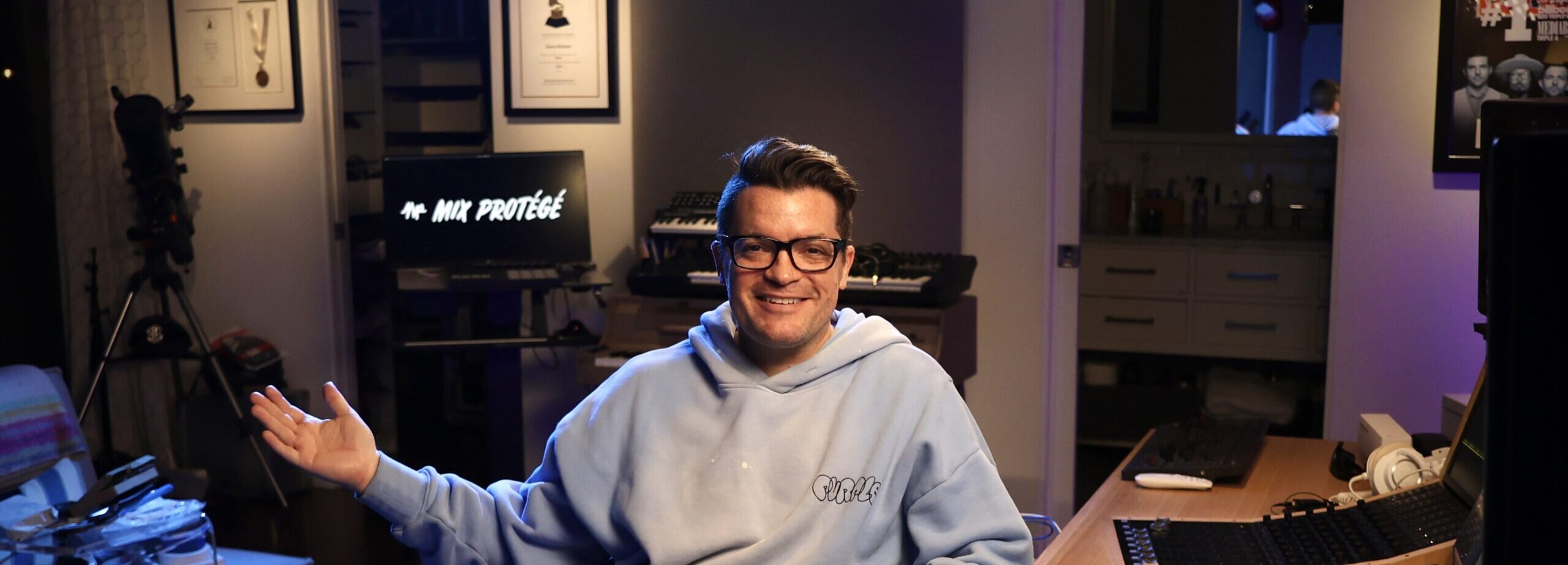 Person seated in a home studio, smiling with one arm raised, surrounded by mixing consoles and keyboards; monitor displays ‘Mix Protégé.