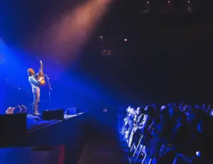 Langhorne Slim performing live onstage, lifting an acoustic guitar toward the lights as the crowd watches from the front barrier.