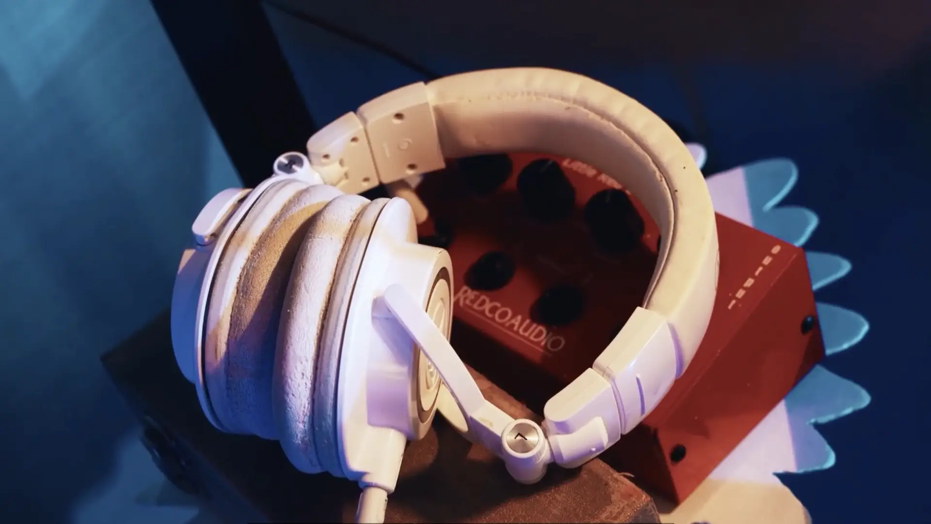 White over-ear headphones resting on a red audio interface with multiple knobs; studio lighting casts blue and warm tones.
